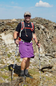 Man hiking in the mountains with trekking poles and wearing a hiking skirt for men.